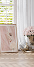 a framed artwork of a woman's face with a pink background, placed on a wooden surface next to a vase of pink roses and a gold tray with a pineapple.